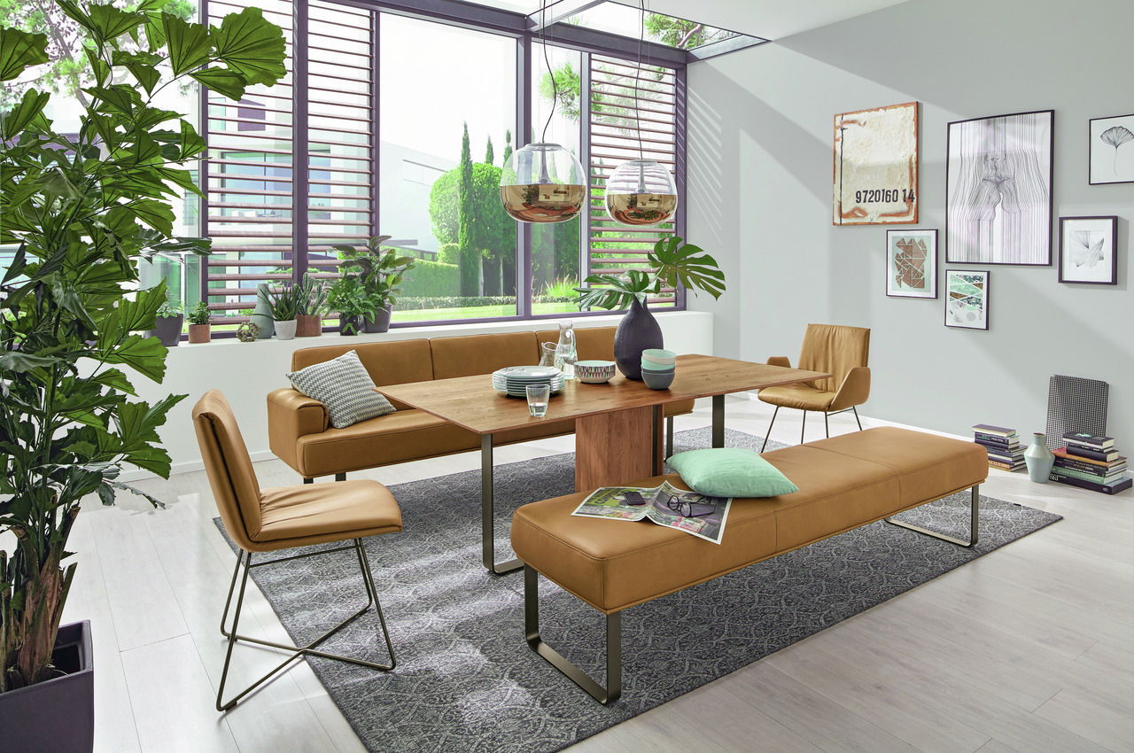 Moderner Esstisch aus Holz mit braunen Lederstühlen und -bänken in einem hellen, stilvoll eingerichteten Raum mit großen Fenstern und Pflanzen, seitliche Perspektive.