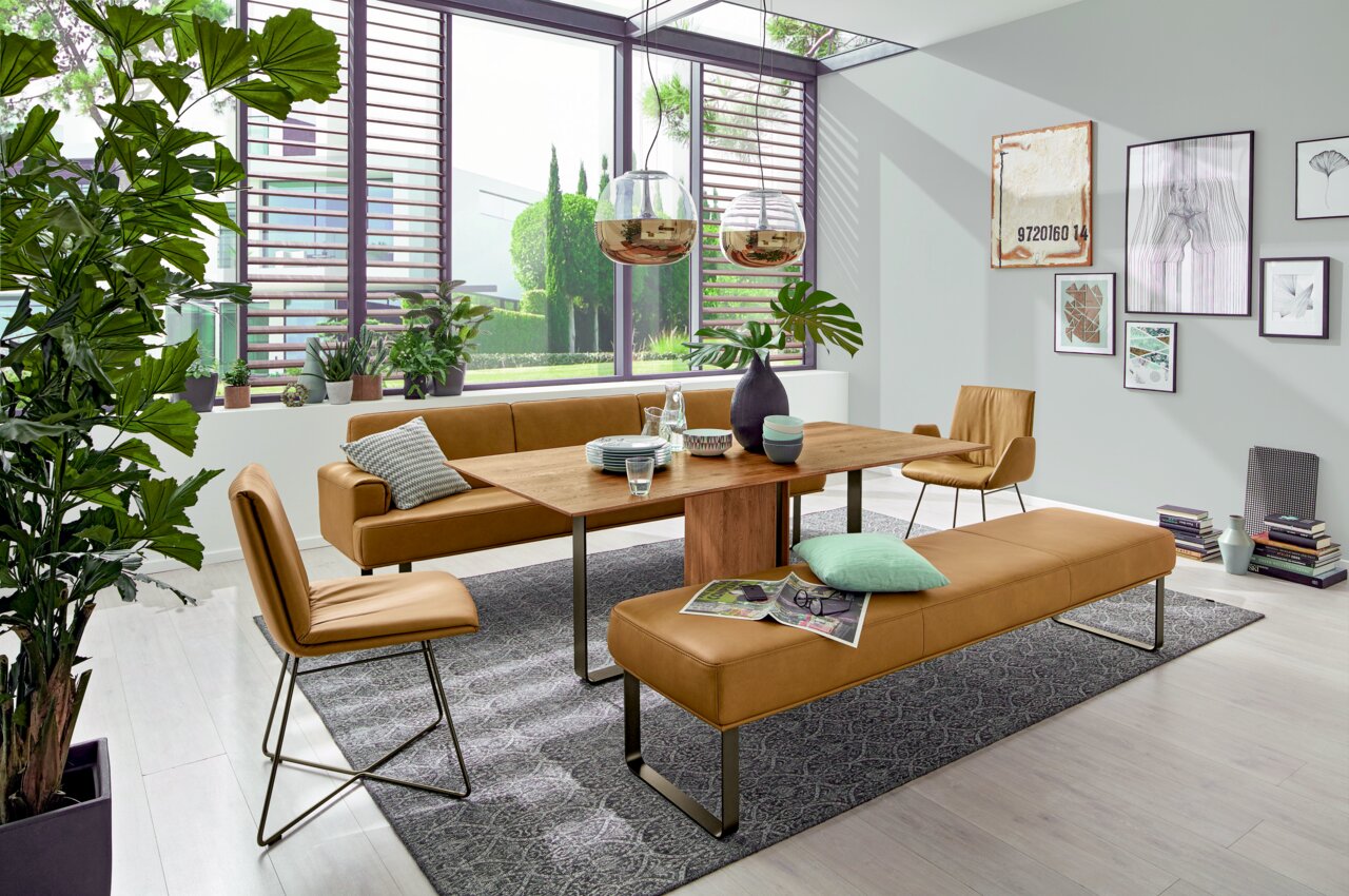 Moderner Esstisch aus Holz mit braunen Lederstühlen und -bänken in einem hellen, stilvoll eingerichteten Raum mit großen Fenstern und Pflanzen, seitliche Perspektive.