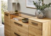 Holz-Kommode aus Eiche mit Schubladen und mittigem offenen Fach mit dunkler Glasablage, dekoriert mit Tischlampe, Büchern und Vase – Ansicht schräg von vorne (leicht von oben).