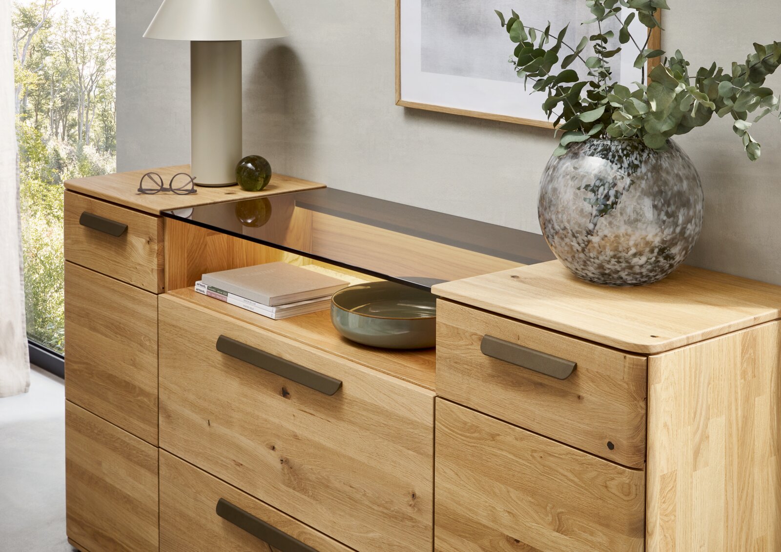 Holz-Kommode aus Eiche mit Schubladen und mittigem offenen Fach mit dunkler Glasablage, dekoriert mit Tischlampe, Büchern und Vase – Ansicht schräg von vorne (leicht von oben).