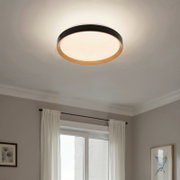 Runde, flache LED-Deckenleuchte mit schwarzem Rand und Holzoptik-Ring, eingeschaltet an weißer Zimmerdecke, fotografiert aus leichter Untersicht in einem Wohnraum.
