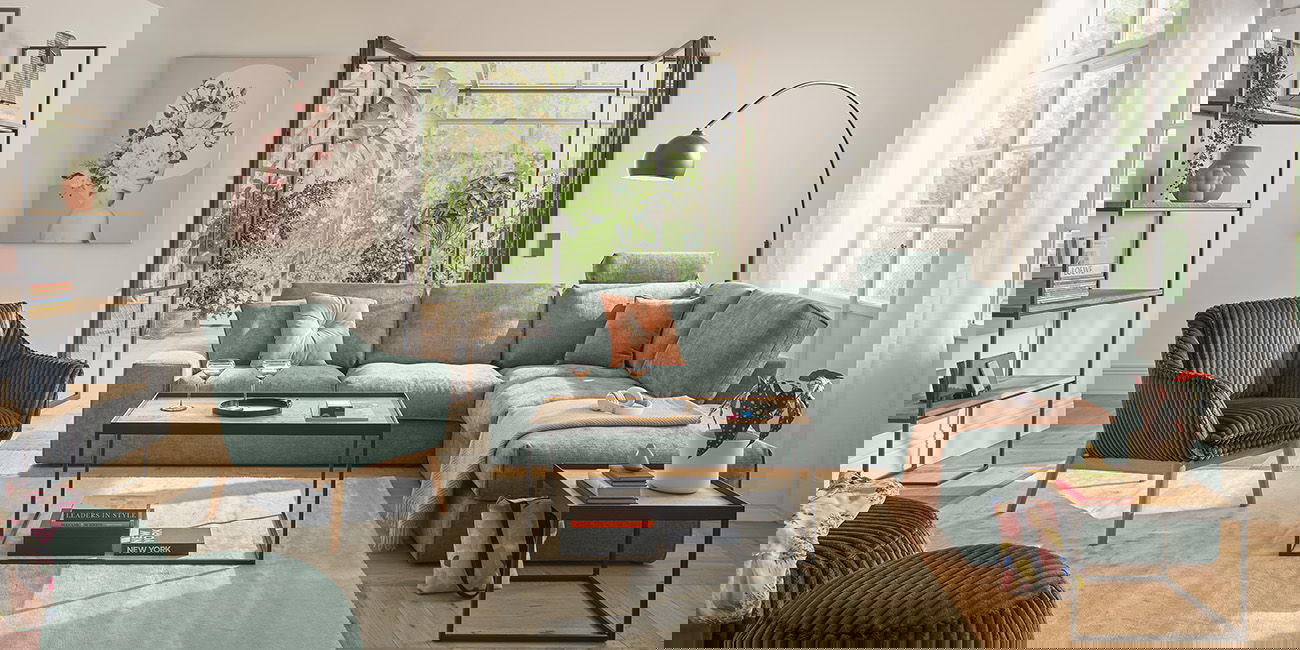 Ein helles WEin Wohnzimmer mit einem großen grünen Sofa und einem passenden Sessel.ohnzimmer mit einem großen grünen Sofa und einem passenden Sessel. Zwei kleine Tische stehen auf einem hellen Teppich. Durch die Glastüren sieht man viele Pflanzen.