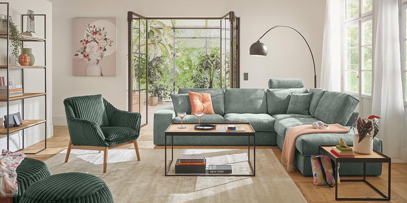 Ein helles WEin Wohnzimmer mit einem großen grünen Sofa und einem passenden Sessel.ohnzimmer mit einem großen grünen Sofa und einem passenden Sessel. Zwei kleine Tische stehen auf einem hellen Teppich. Durch die Glastüren sieht man viele Pflanzen.