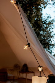 Schräg von unten aufgenommene Nahaufnahme einer warm leuchtenden Lichterkette am Rand eines beigen Glamping-Zelts, mit unscharfem Innenraum und Bäumen im Hintergrund.