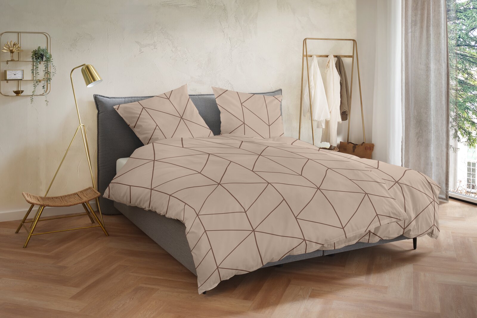 Beige Bettwäsche mit geometrischem Linienmuster in Braun auf einem grauen Polsterbett, frontal in leichter Schrägansicht von vorn fotografiert, im hellen Schlafzimmer-Setting mit goldener Stehlampe und Kleiderständer.