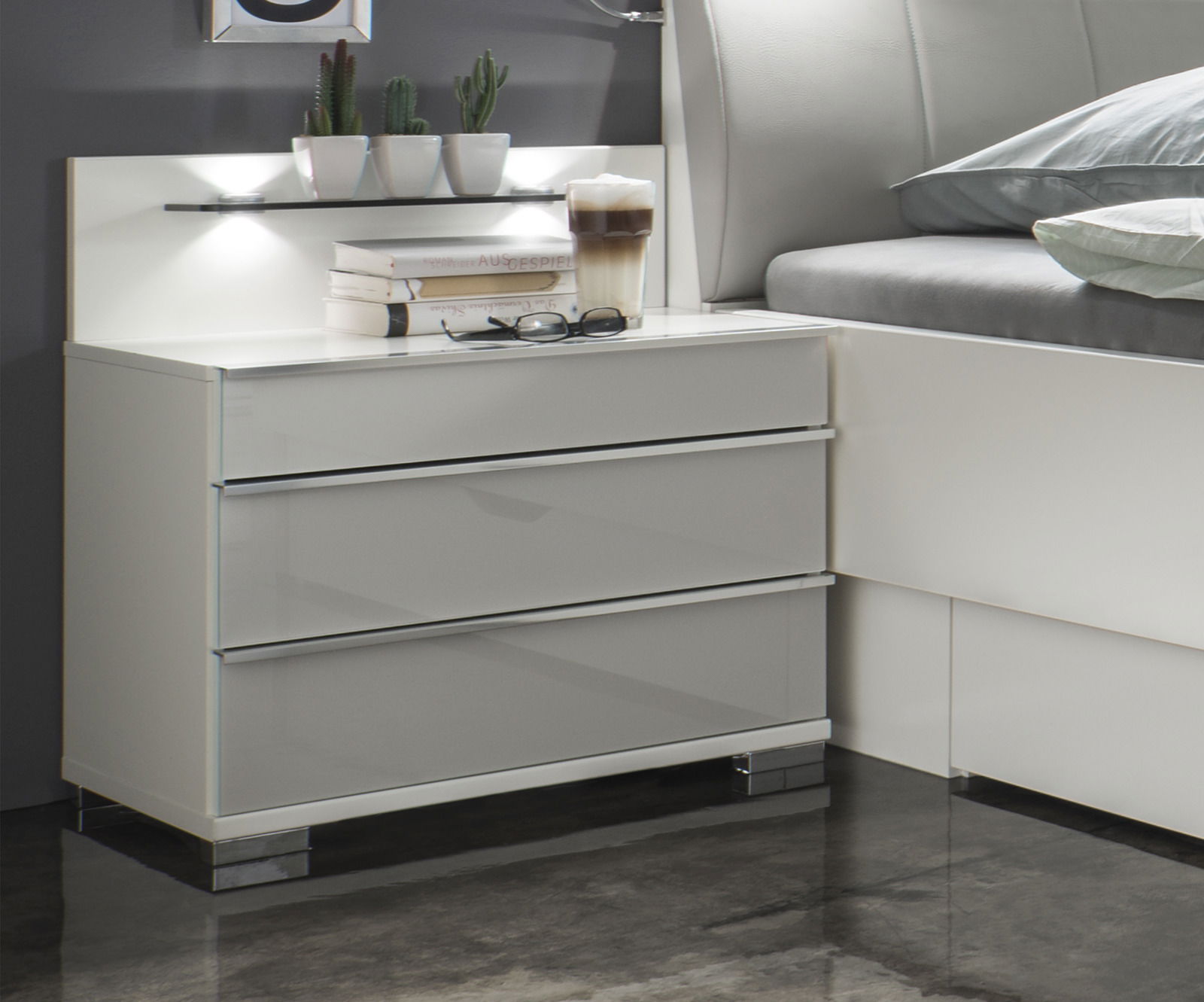 Moderne Nachtkonsole mit drei Schubladen in Hochglanzoptik, seitliche Perspektive, dekoriert mit Büchern, Brille und Kaktuspflanzen.