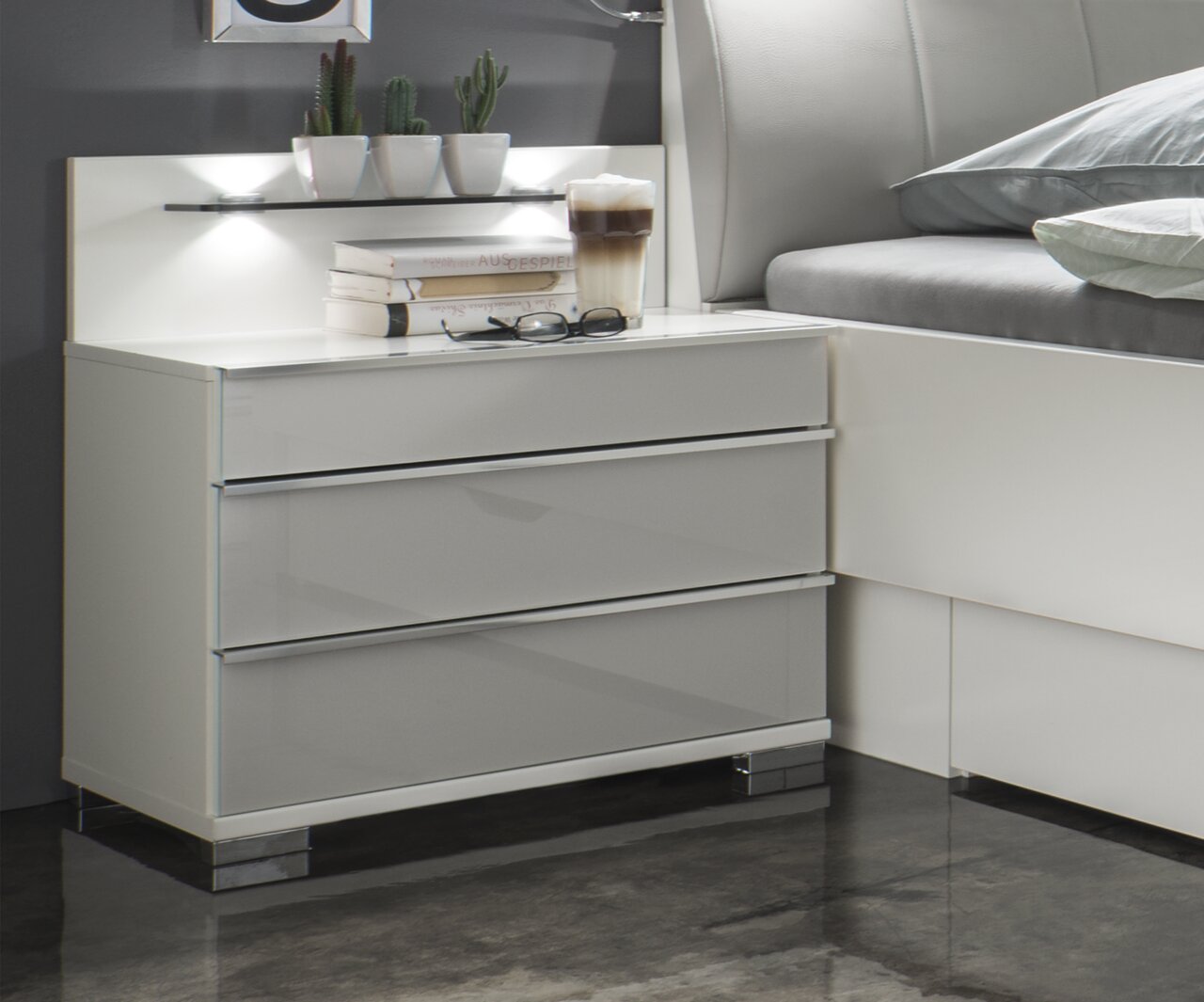 Novel Nachtkonsolen-Paar SHANGHAI 2 Moderne Nachtkonsole mit drei Schubladen in Hochglanzoptik, seitliche Perspektive, dekoriert mit Büchern, Brille und Kaktuspflanzen.