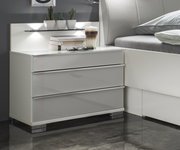 Moderne Nachtkonsole mit drei Schubladen in Hochglanzoptik, seitliche Perspektive, dekoriert mit Büchern, Brille und Kaktuspflanzen.