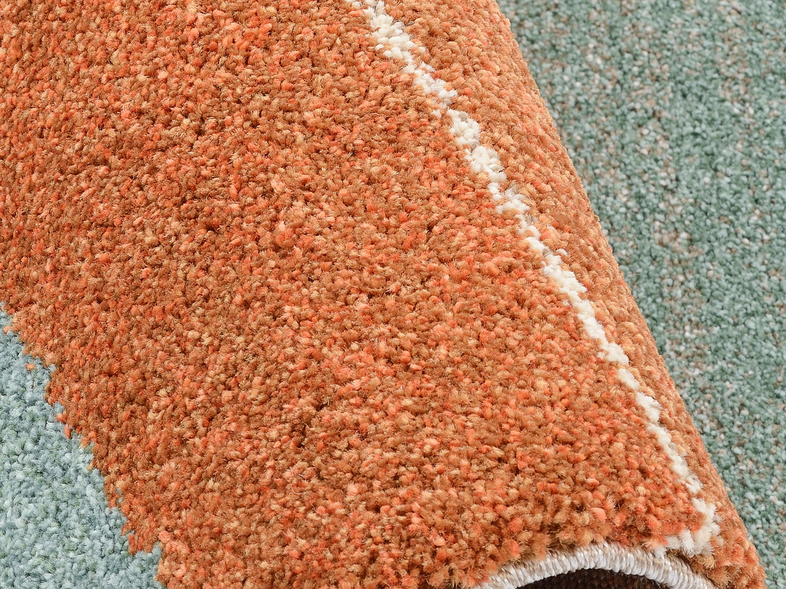Nahaufnahme eines orangefarbenen Frisé-Teppichs mit einer weißen Linie, die diagonal verläuft. Der Teppich ist teilweise aufgerollt und zeigt eine weiche, flauschige Textur.