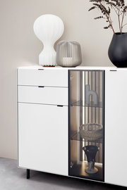 Modernes Highboard mit weißer Oberfläche, zwei Schubladen und einer Glastür, dekoriert mit einer weißen Lampe, einer grauen Laterne und einer schwarzen Vase. Frontale Perspektive.