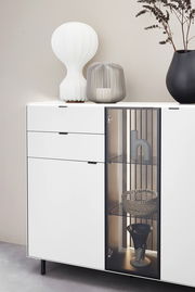 Modernes Highboard mit weißer Oberfläche, zwei Schubladen und einer Glastür, dekoriert mit einer weißen Lampe, einer grauen Laterne und einer schwarzen Vase. Frontale Perspektive.