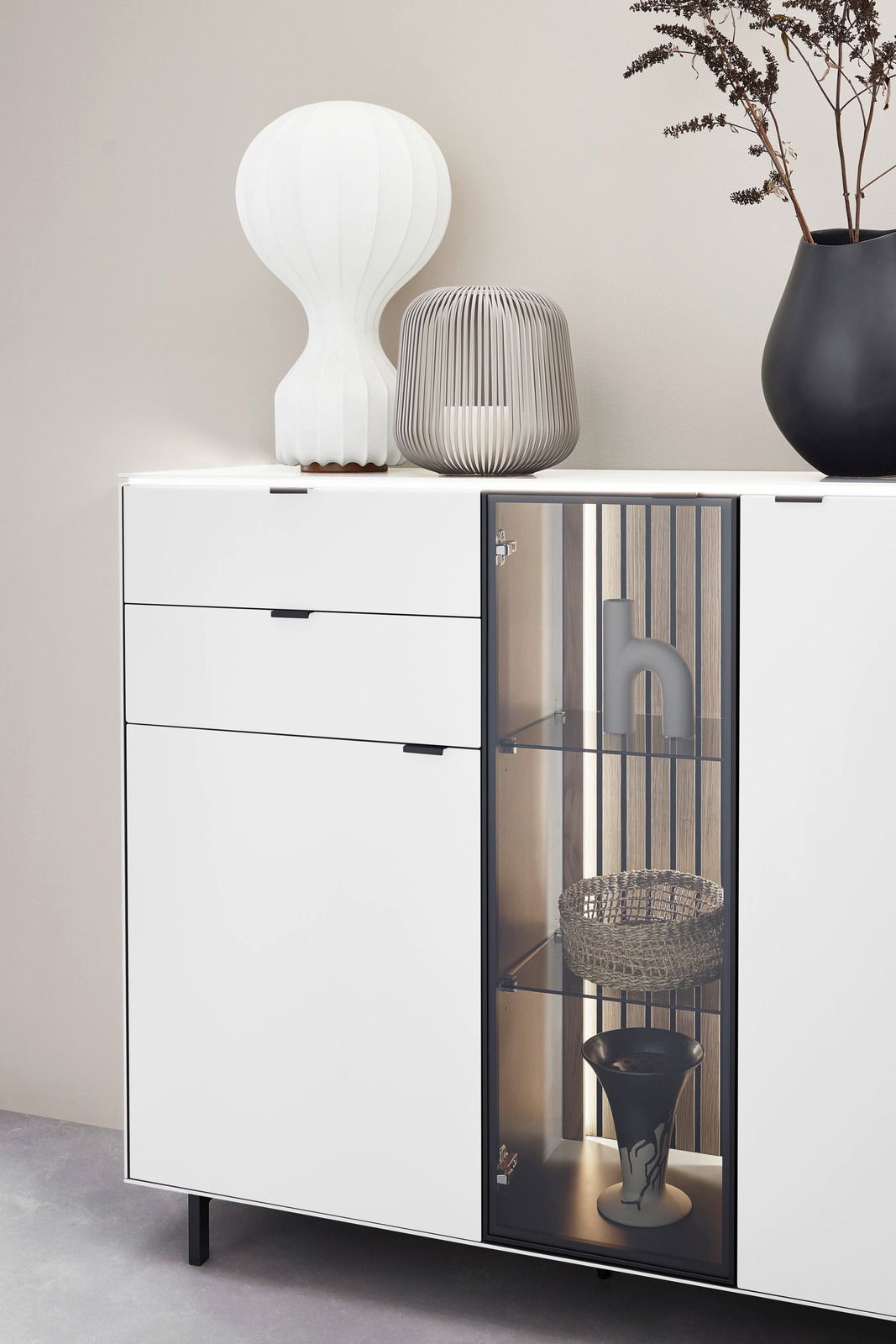 Modernes Highboard mit weißer Oberfläche, zwei Schubladen und einer Glastür, dekoriert mit einer weißen Lampe, einer grauen Laterne und einer schwarzen Vase. Frontale Perspektive.