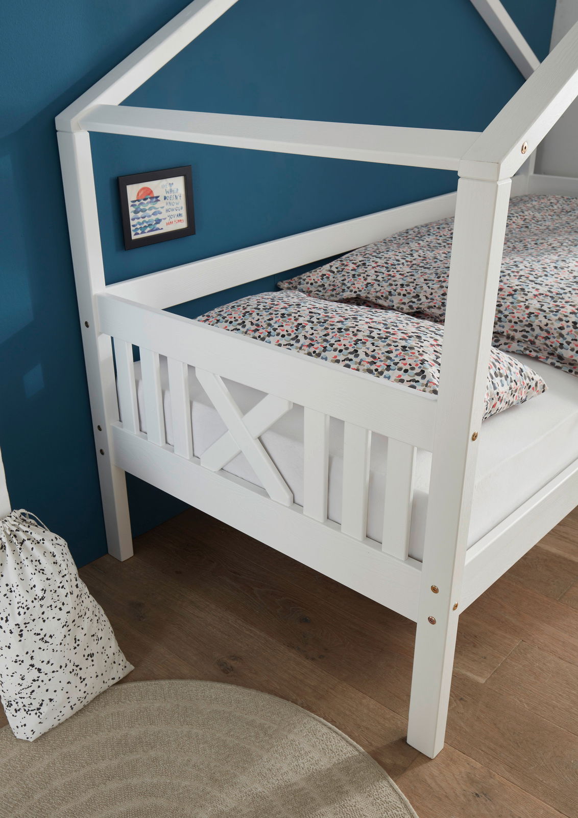 Weißes Hausbett mit Gitterdesign, seitliche Perspektive, vor einer blauen Wand mit gemusterten Bettbezügen.