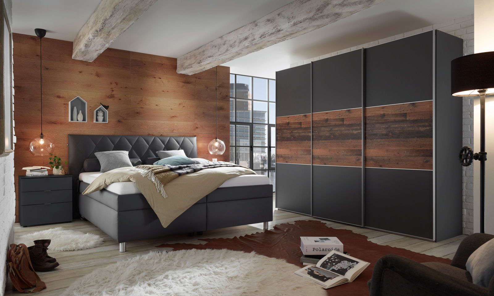 Modernes Schlafzimmer mit einem eleganten Polsterbett in dunklem Grau, frontal aufgenommen. Das Bett hat ein gestepptes Kopfteil und ist mit neutralen Kissen und Decken dekoriert. Im Hintergrund sind ein großer Kleiderschrank mit Holzelementen und eine Ho