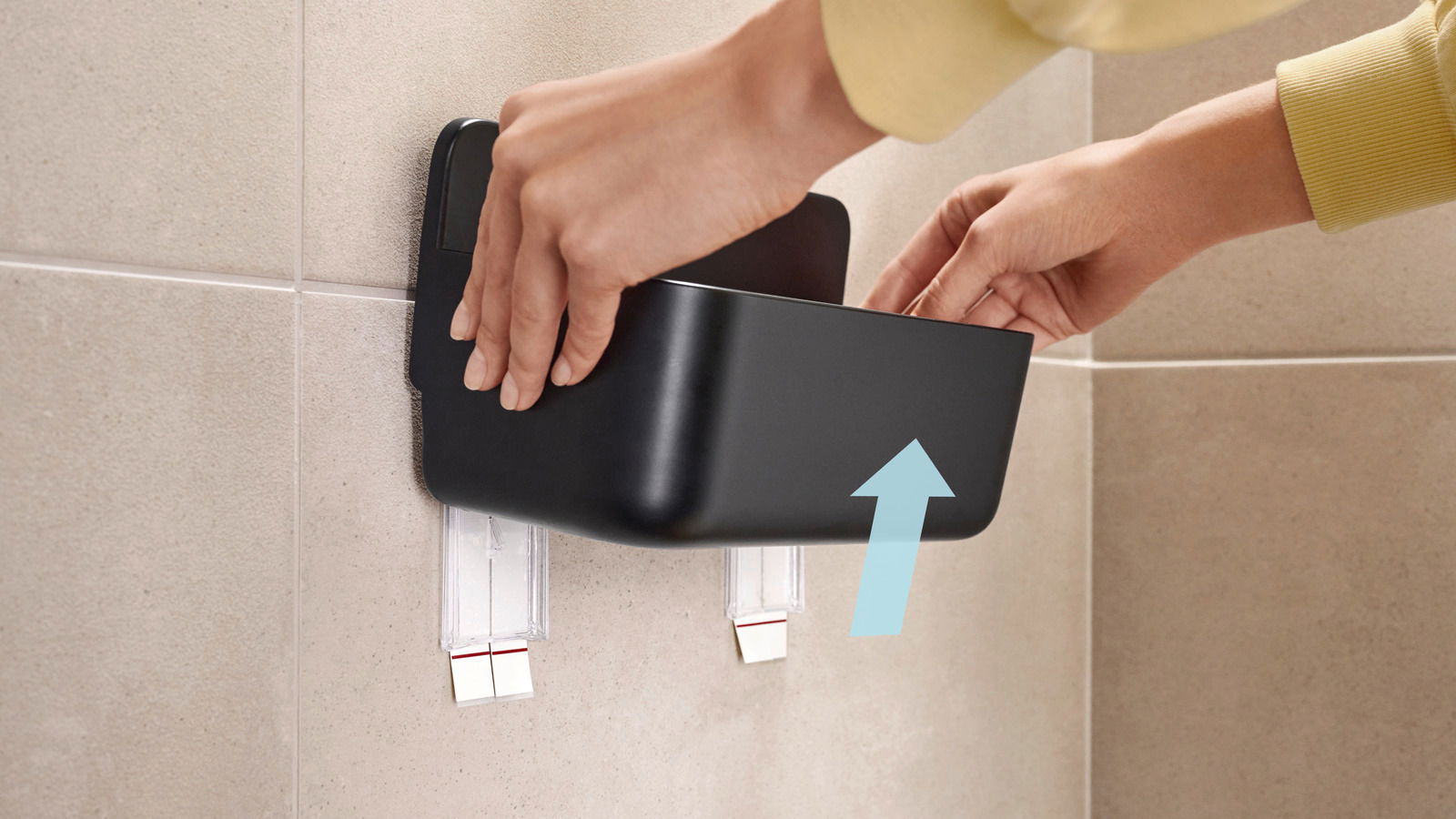 Eine Person montiert einen schwarzen Duschkorb an einer gefliesten Badezimmerwand. Der Korb wird von zwei transparenten Klebestreifen gehalten. Perspektive von der Seite.