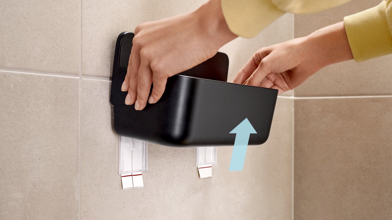 Eine Person montiert einen schwarzen Duschkorb an einer gefliesten Badezimmerwand. Der Korb wird von zwei transparenten Klebestreifen gehalten. Perspektive von der Seite.