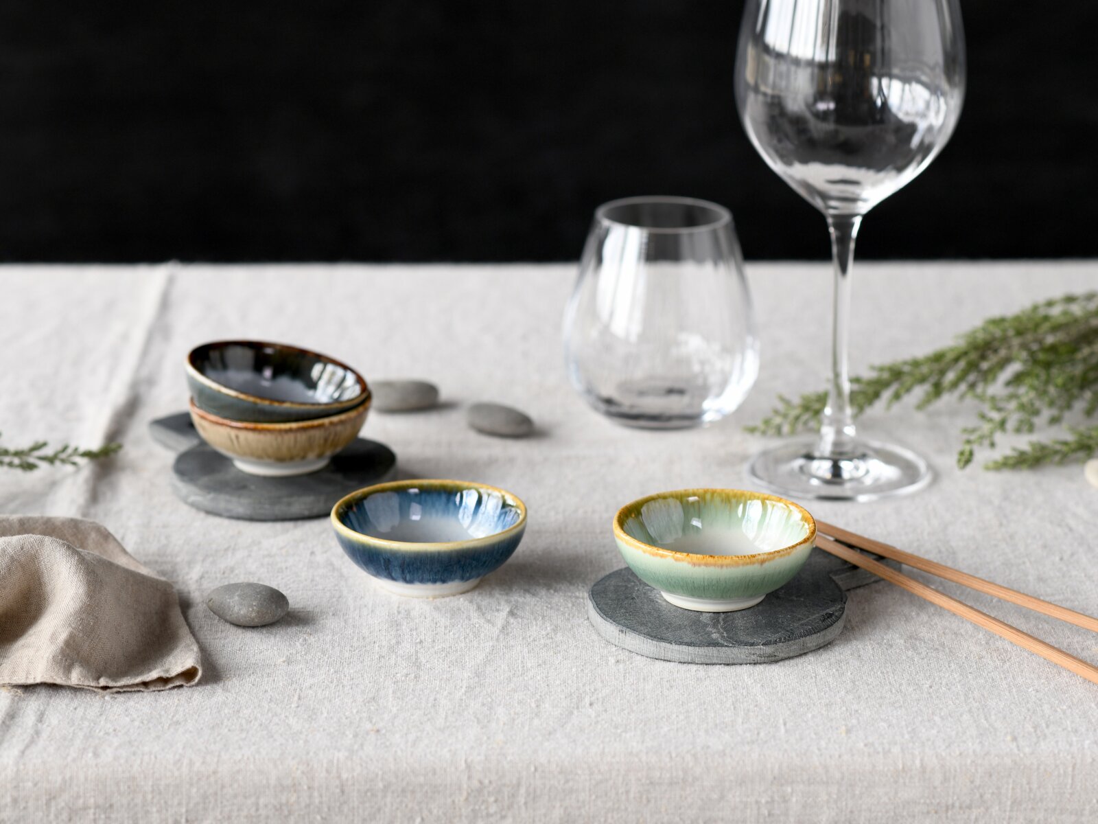 Drei kleine glasierte Keramikschalen in Blau, Grün und Beige auf grauer Tischdecke mit Schieferuntersetzern, Essstäbchen und Weingläsern, aufgenommen aus leicht erhöhter Frontperspektive