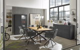Moderner Esstisch mit Holzplatte und schwarzen Metallbeinen, umgeben von grauen Stühlen in einem stilvollen Esszimmer mit großen Fenstern und urbanem Ausblick, seitliche Perspektive.