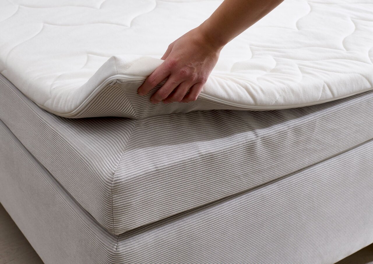 Close-up van een boxspringbed met een hand die het bovenste matras optilt om de constructie en het comfort te laten zien.