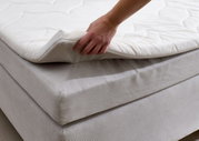 Close-up van een boxspringbed met een hand die het bovenste matras optilt om de constructie en het comfort te laten zien.
