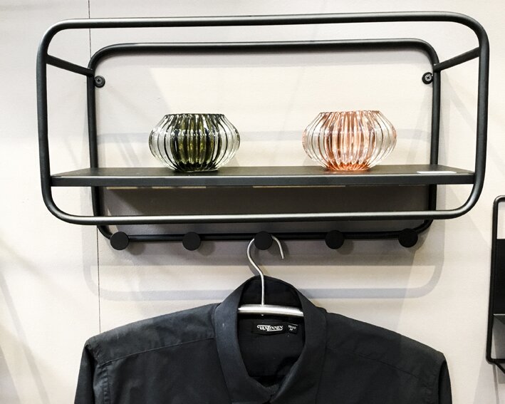 Zurbrüggen Wandgarderobe Schwarze Wandgarderobe aus Metall mit Ablage und fünf Haken, frontal aus leichter Untersicht fotografiert; darüber stehen zwei gerippte Glas-Teelichthalter, darunter hängt eine schwarze Jacke am Kleiderbügel.