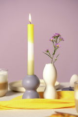 Keramikleuchter in Fliederfarbe mit brennender Kerze, seitliche Perspektive, auf einem Tisch mit gelbem Tuch und Vase mit lila Blumen.