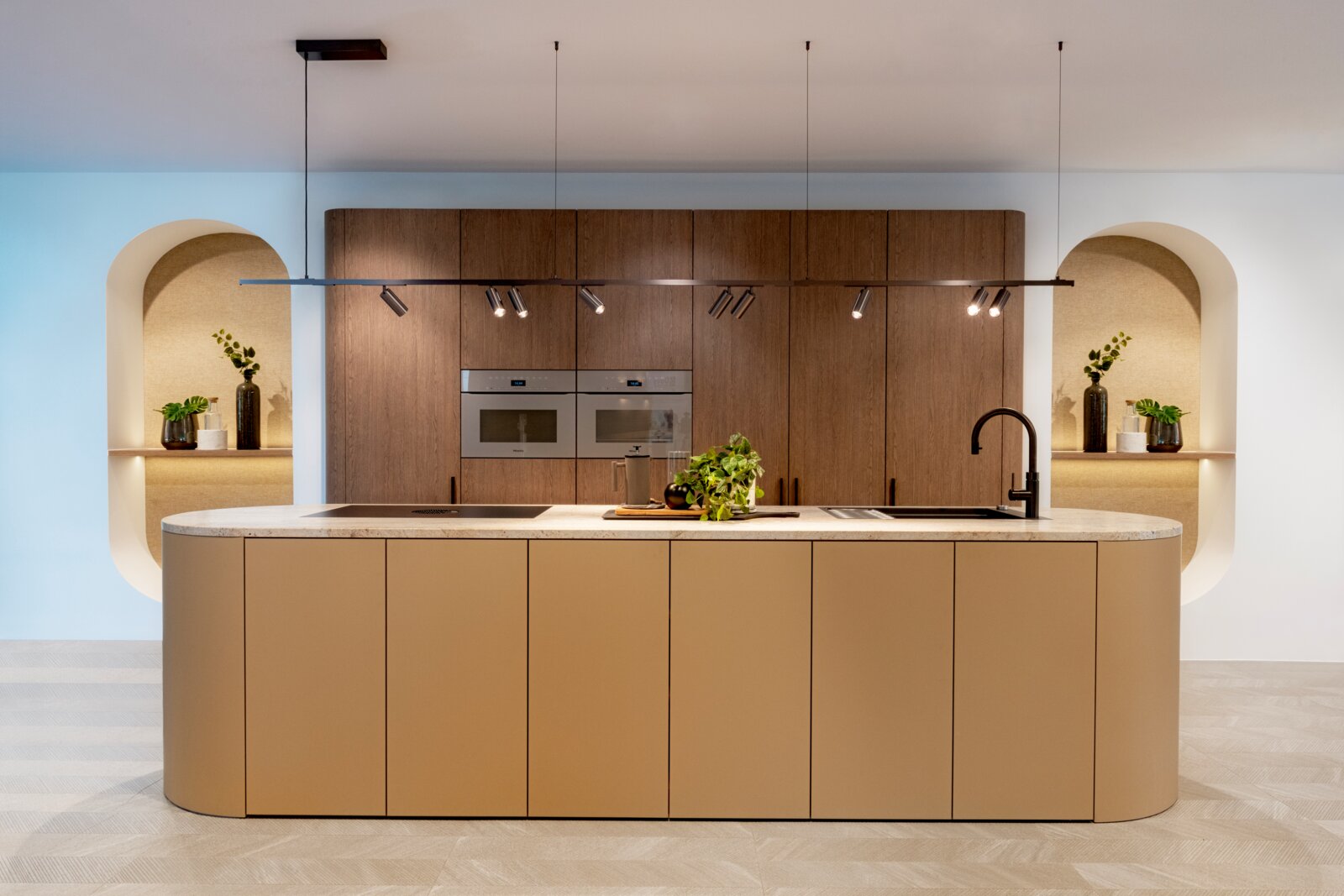 Moderne Designküche mit großer Kücheninsel in Beige und heller Stein-Arbeitsplatte, Holzfronten mit Einbauöfen, schwarzer Armatur und Hängeleuchte; frontale Perspektive auf die Küchenzeile.
