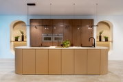 Moderne Designküche mit großer Kücheninsel in Beige und heller Stein-Arbeitsplatte, Holzfronten mit Einbauöfen, schwarzer Armatur und Hängeleuchte; frontale Perspektive auf die Küchenzeile.