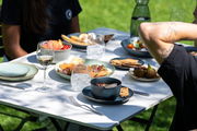 Müslischale 15 cm auf einem gedeckten Tisch im Freien, gefüllt mit Müsli und Obst, umgeben von weiteren Tellern und Gläsern, seitliche Perspektive.