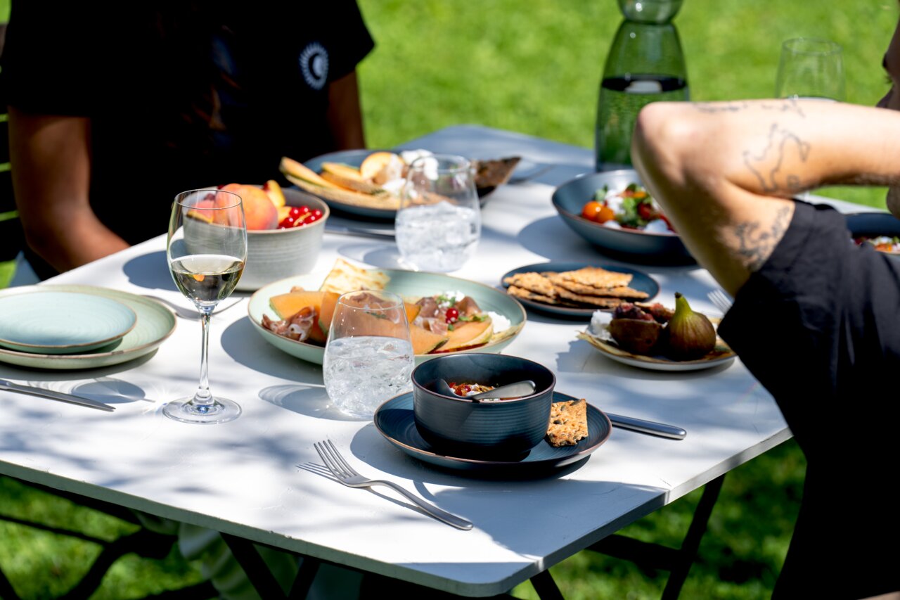 Een mueslischaal van 15 cm op een gedekte tafel in de open lucht, gevuld met muesli en fruit, omringd door andere borden en glazen, zijaanzicht.