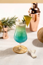 Vooraanzicht van een tropische blauwgroene cocktail met romige schuimlaag en ananasgarnering in een elegant hurricane-glas, omringd door cocktailaccessoires en vers fruit.