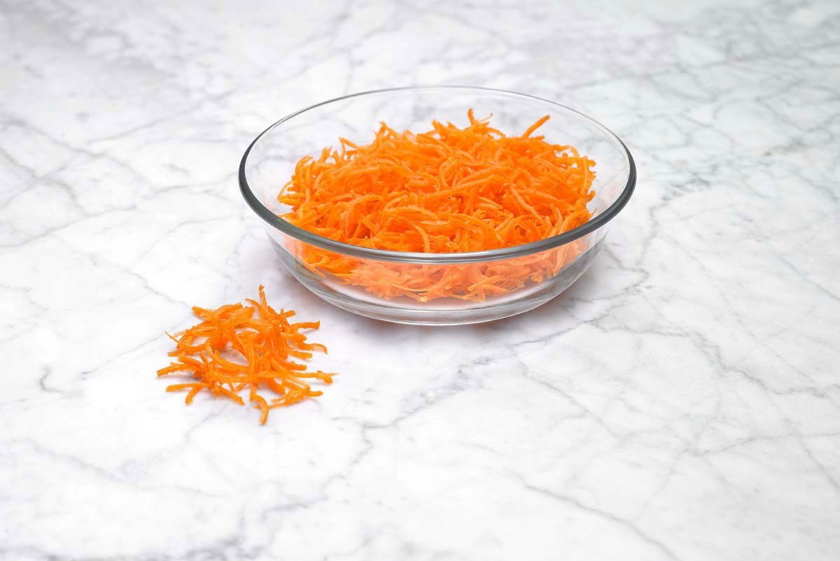 Eine Glasschüssel mit frisch geraspelten Karotten auf einer Marmoroberfläche, seitliche Perspektive.