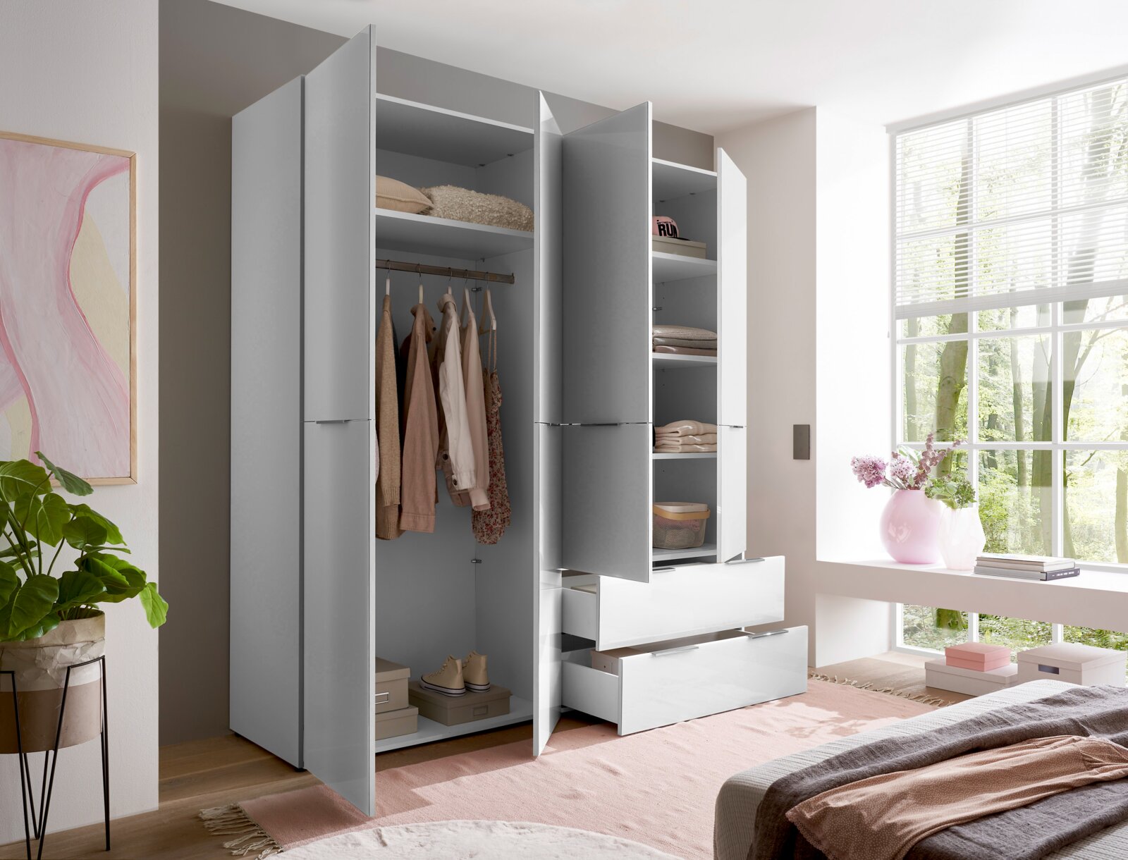 Moderner Kleiderschrank in Grau und Weiß mit geöffneten Türen und zwei ausgezogenen Schubladen, sichtbar sind Kleiderstange und Ablagefächer; Ansicht schräg von vorne im Schlafzimmer neben großem Fenster.