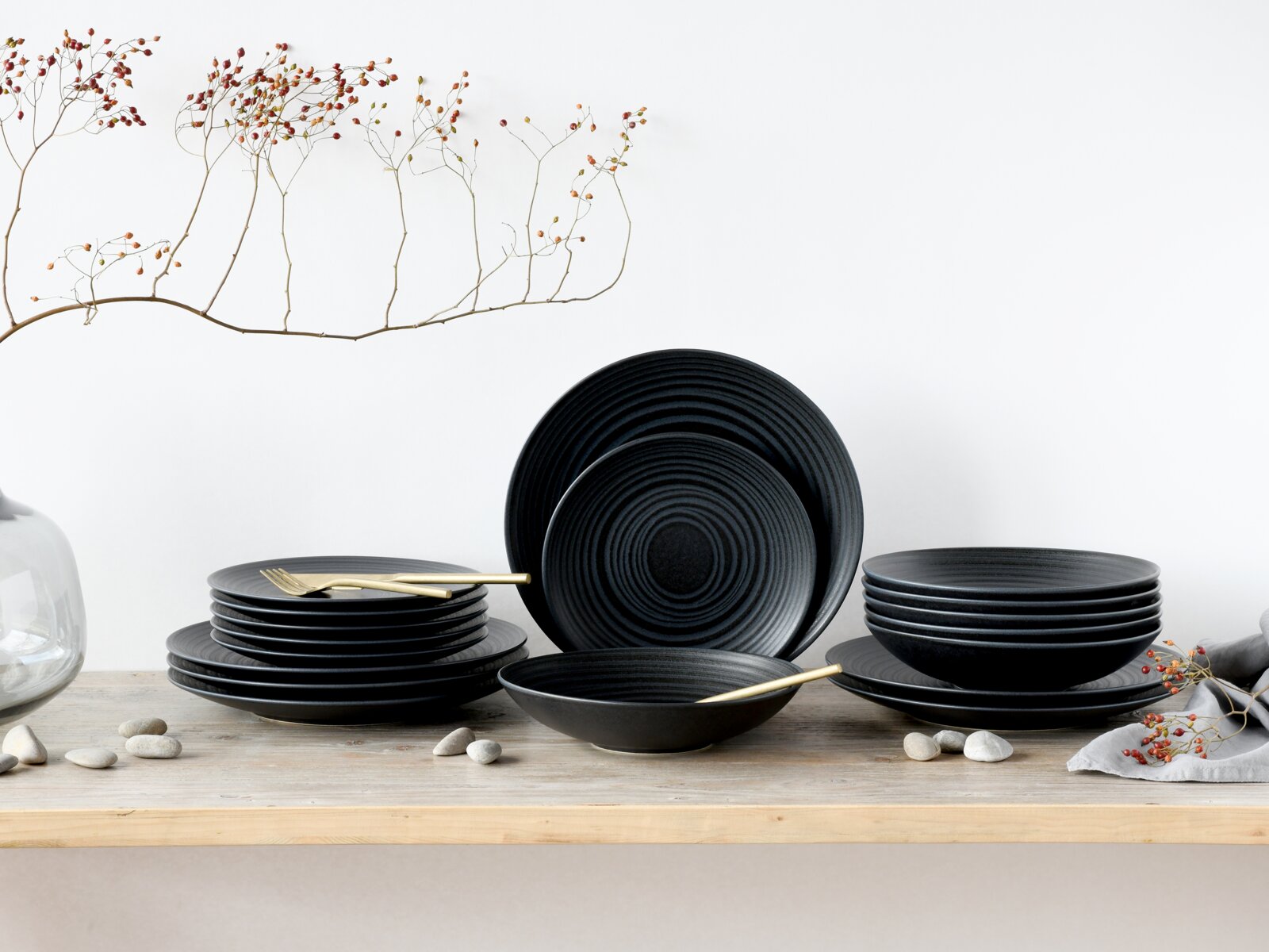 Creatable Tellerset 18-tlg LAVA STONE Schwarzes Geschirrset mit strukturierten Tellern und Schalen auf Holztisch, mit goldfarbenem Besteck und dekorativen Zweigen, Frontansicht auf weißem Hintergrund