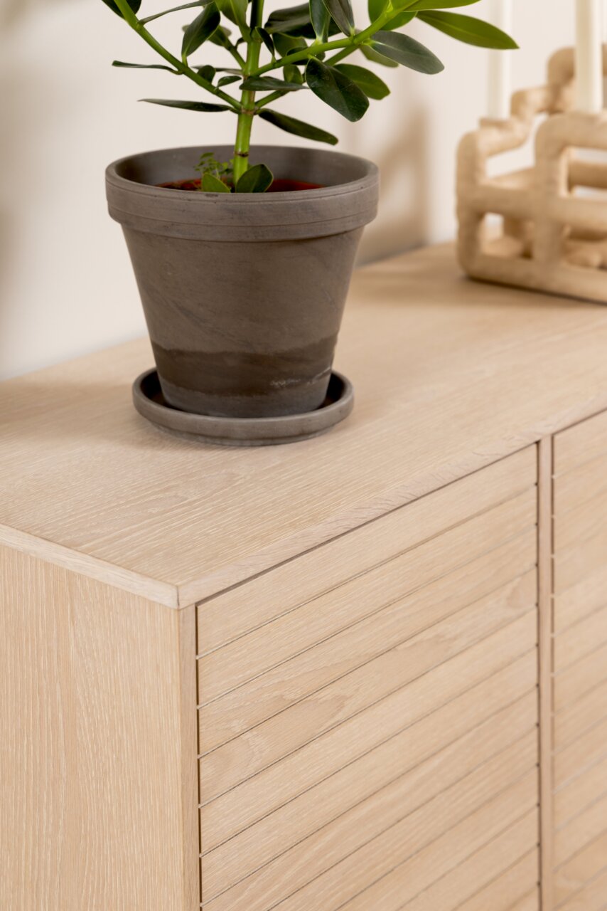 Close-up van een dressoir van licht hout met daarop een plant in een grijze pot, gezien vanuit de zijkant.