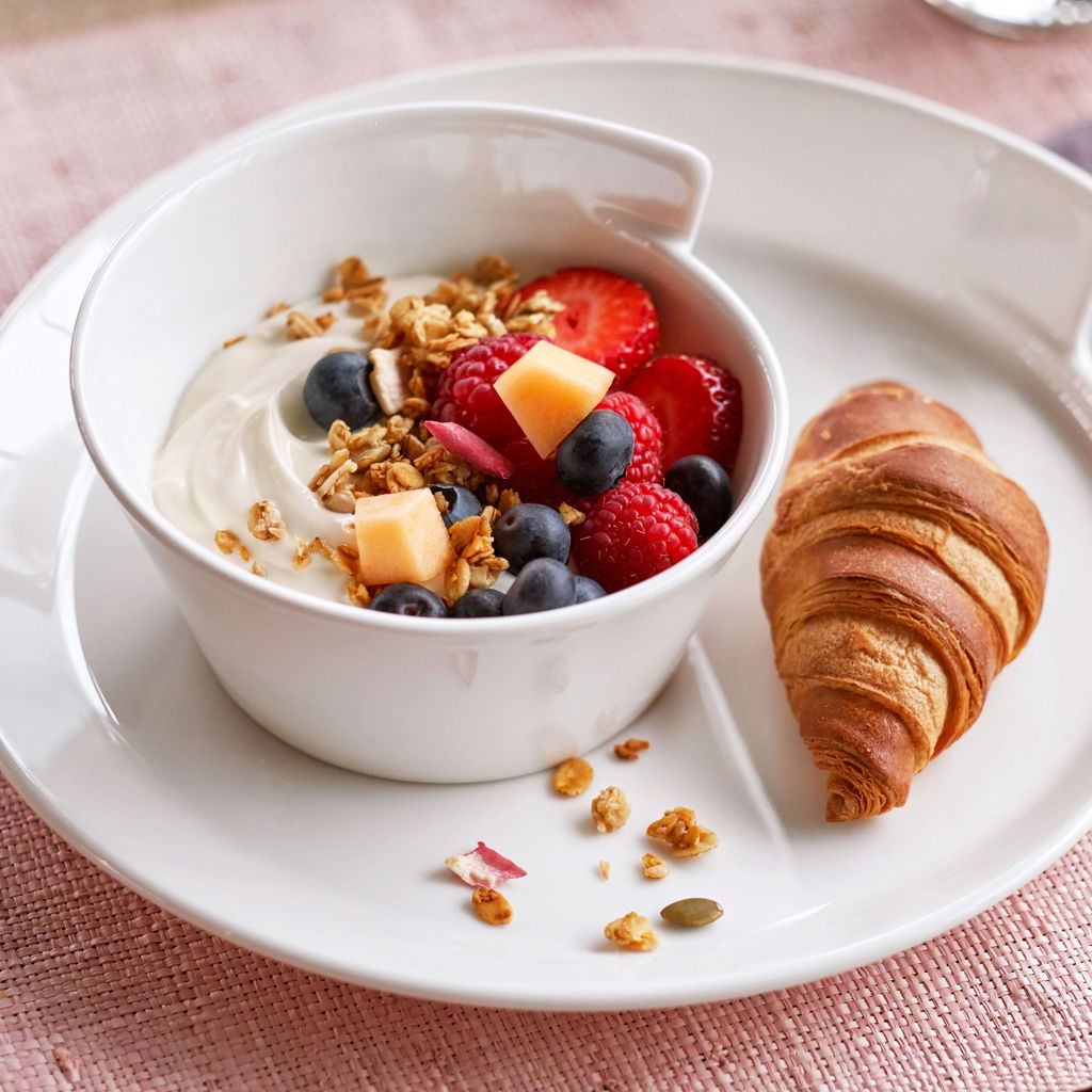 Weiße Schale der Serie NewWave mit Joghurt, frischen Beeren und Müsli, daneben ein Croissant auf einem Teller, aus der Vogelperspektive fotografiert.