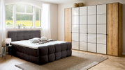 Modernes Boxbett in einem hellen Schlafzimmer mit großem Fenster, grauem gepolstertem Kopfteil und passendem Schrank, seitliche Perspektive.