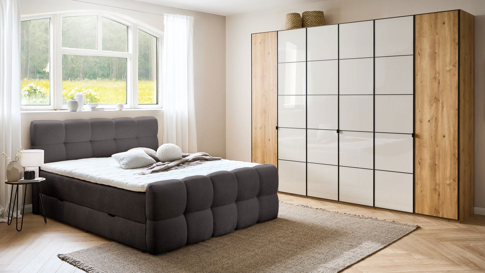 Modernes Boxbett in einem hellen Schlafzimmer mit großem Fenster, grauem gepolstertem Kopfteil und passendem Schrank, seitliche Perspektive.