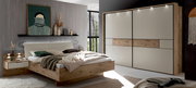Modernes Schlafzimmer-Set mit Holzbett und gepolstertem Kopfteil, passendem Nachttisch und großem Schwebetürenschrank in Weiß mit Holz-Akzent und LED-Spots, fotografiert in schräger Frontansicht aus dem Raum.