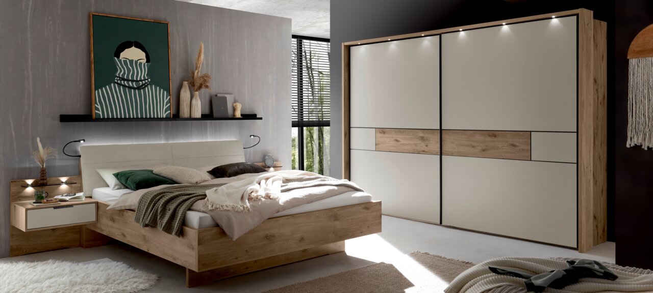 Modernes Schlafzimmer-Set mit Holzbett und gepolstertem Kopfteil, passendem Nachttisch und großem Schwebetürenschrank in Weiß mit Holz-Akzent und LED-Spots, fotografiert in schräger Frontansicht aus dem Raum.