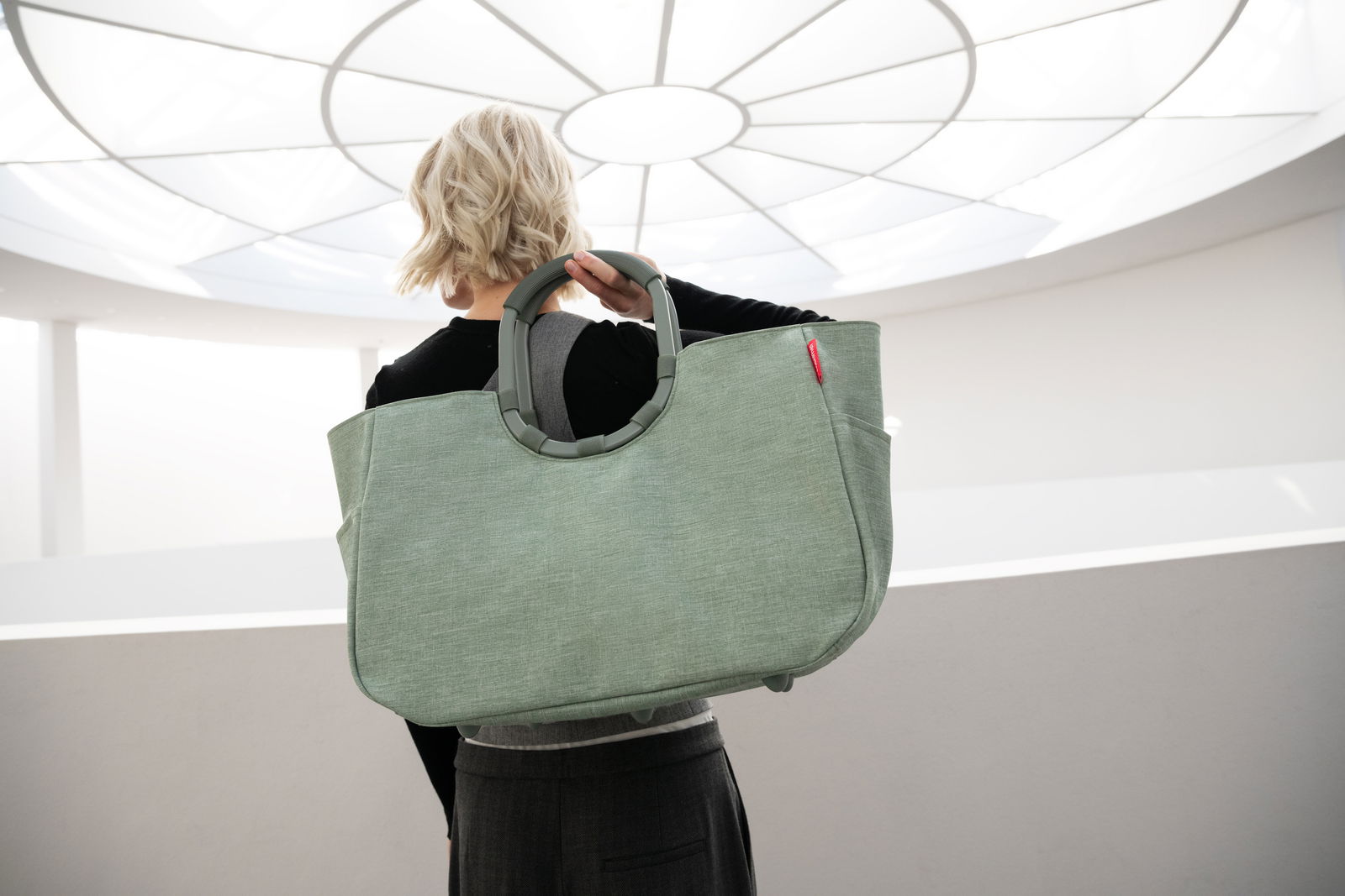 Rückansicht einer Person, die eine große, grüne Tasche mit runden Griffen trägt, in einem hellen, modernen Raum.
