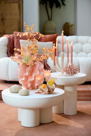Moderner Couchtisch mit runder, weißer Oberfläche, dekoriert mit Kerzen, Muscheln und einer Vase mit orangefarbenen Blumen, aus seitlicher Perspektive fotografiert.