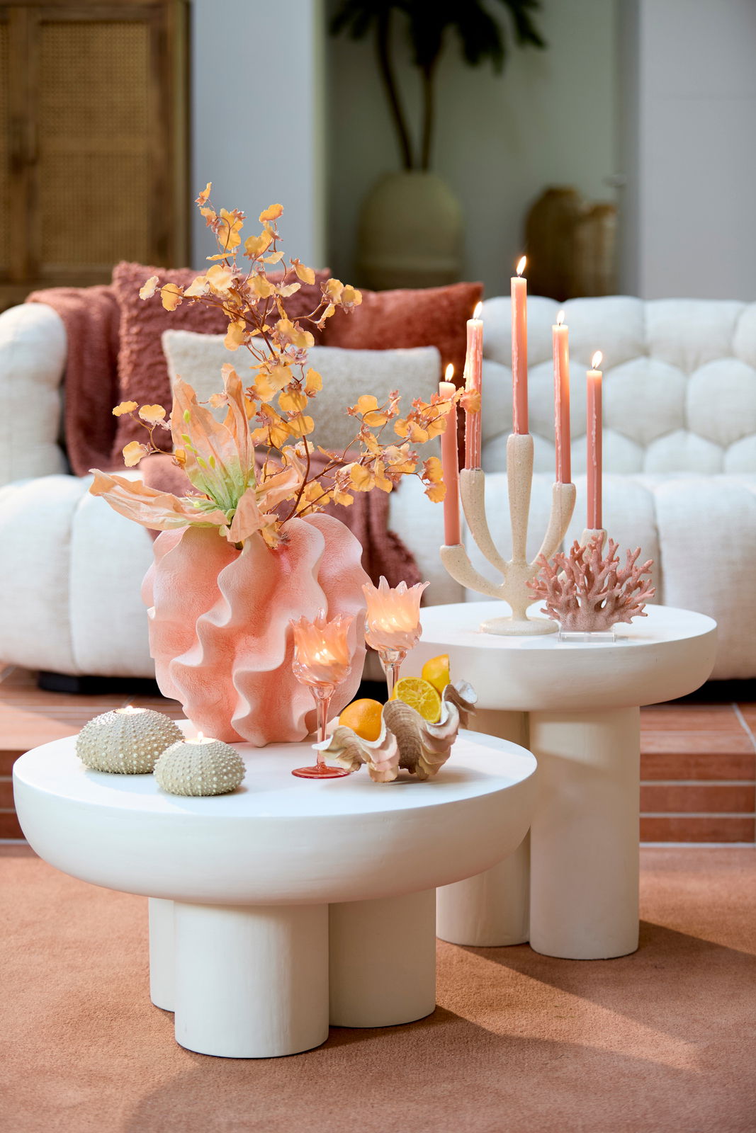 Moderner Couchtisch mit runder, weißer Oberfläche, dekoriert mit Kerzen, Muscheln und einer Vase mit orangefarbenen Blumen, aus seitlicher Perspektive fotografiert.