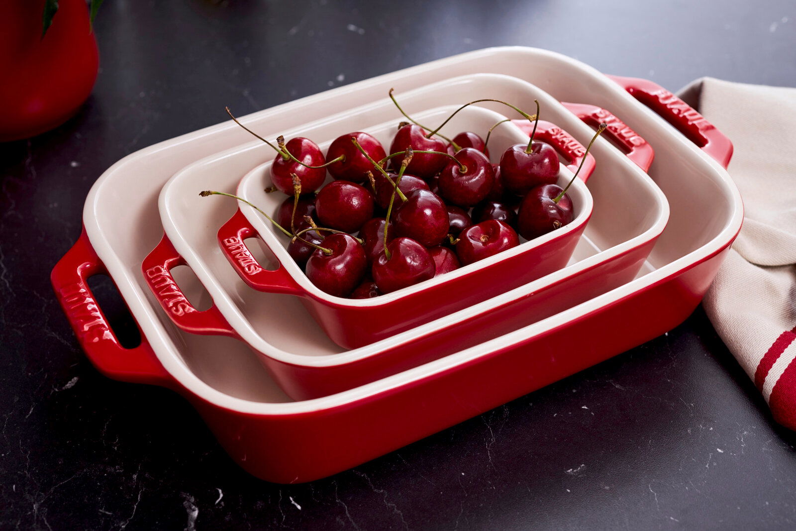 Dreiteiliges Auflaufformenset in Rot, gestapelt und gefüllt mit Kirschen, aus der Vogelperspektive fotografiert.