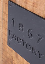 Nahaufnahme eines Highboards mit einer schwarzen Metallplatte, auf der '1867 FACTORY' steht. Die Perspektive zeigt die Platte schräg von der Seite.