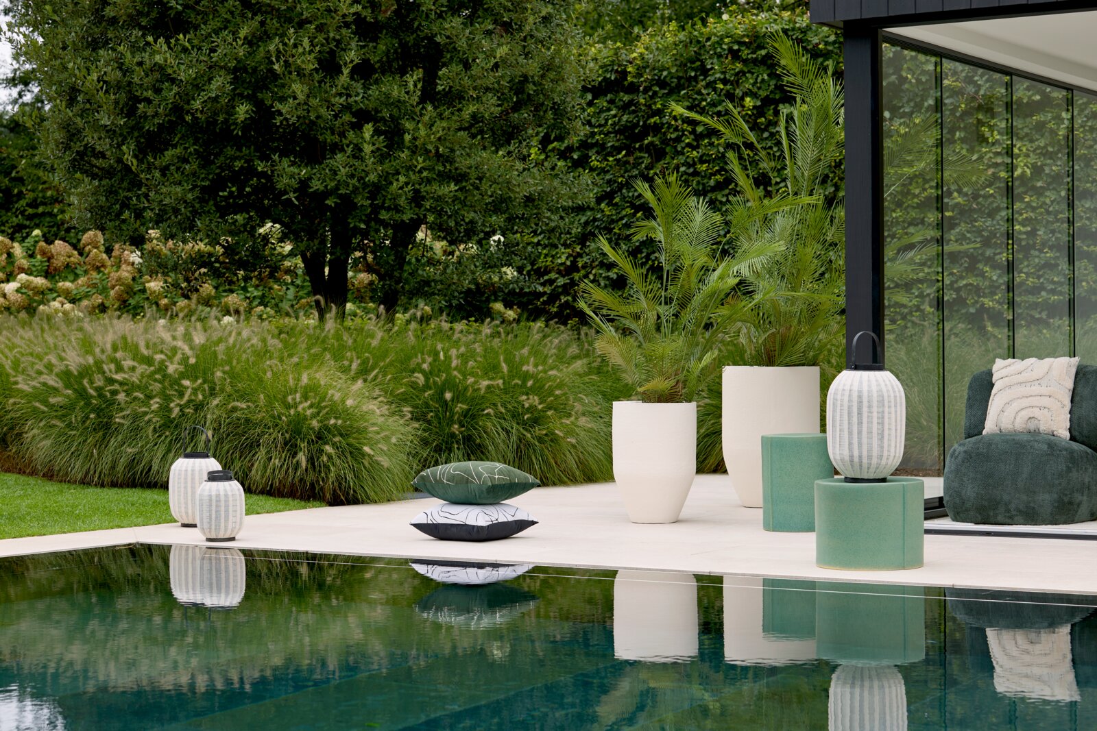Breed frontaal perspectief op een stijlvolle poolside setting met decoratieve lantaarns, groene bijzettafels, moderne plantenbakken en sierkussens in een weelderige tuin.