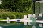 Breed frontaal perspectief op een stijlvolle poolside setting met decoratieve lantaarns, groene bijzettafels, moderne plantenbakken en sierkussens in een weelderige tuin.