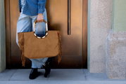 Braune Wildleder-Optik Shopper-Handtasche mit Fransen und rundem Griff, von vorn in leichter Seitenansicht fotografiert, getragen von einer Person vor einer Tür