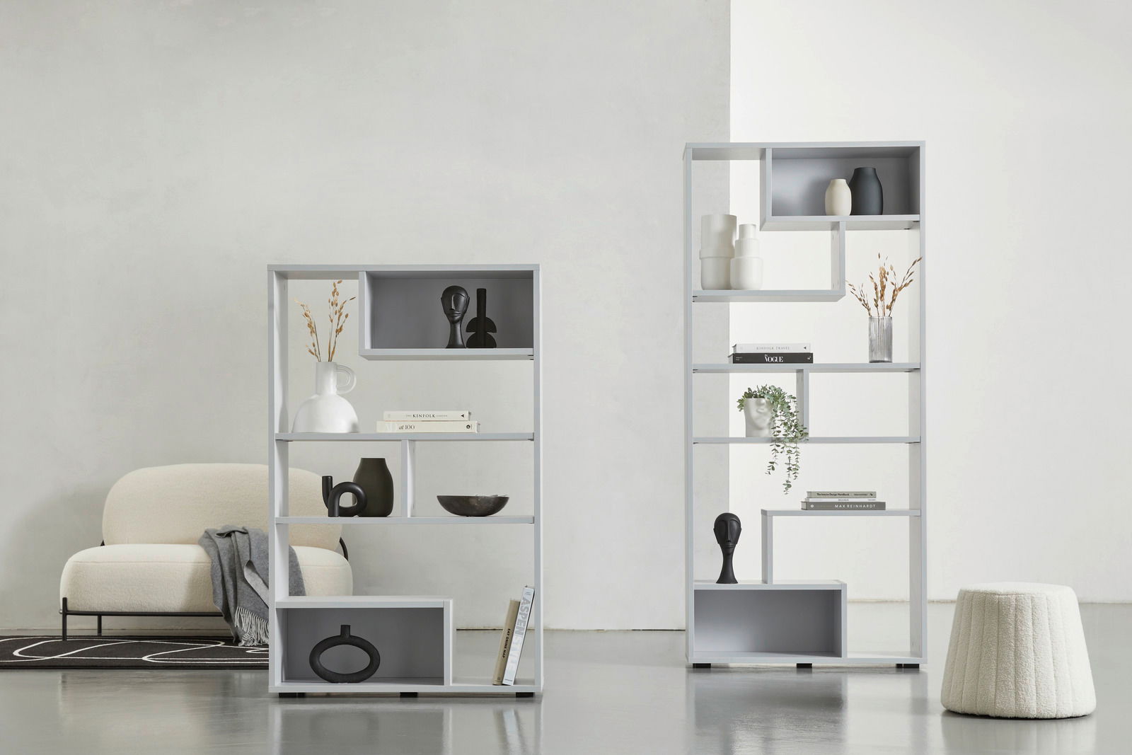 Hom´in Raumteiler hoch JOLLY Zwei moderne, weiße Raumteiler mit dekorativen Objekten, von vorne betrachtet. Der linke Raumteiler ist niedriger und breiter, der rechte ist höher und schmaler. Beide stehen auf einem hellen Boden vor einer weißen Wand.