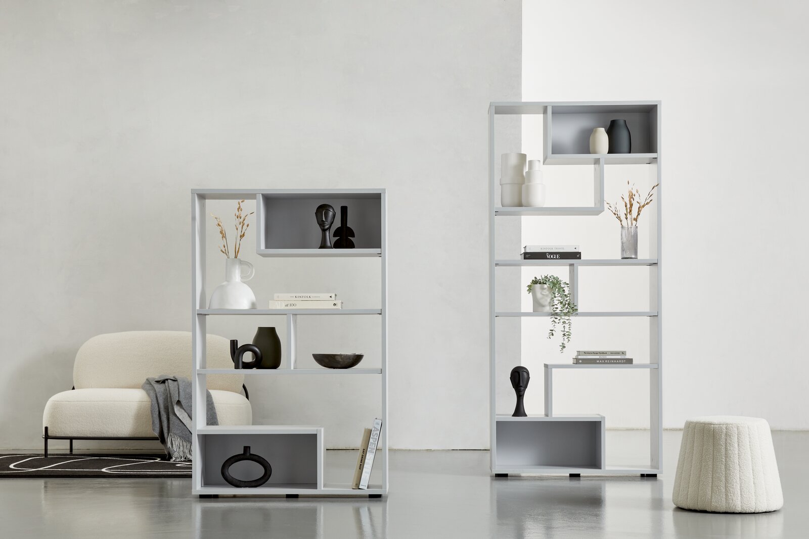 Zwei moderne, weiße Raumteiler mit dekorativen Objekten, von vorne betrachtet. Der linke Raumteiler ist niedriger und breiter, der rechte ist höher und schmaler. Beide stehen auf einem hellen Boden vor einer weißen Wand.